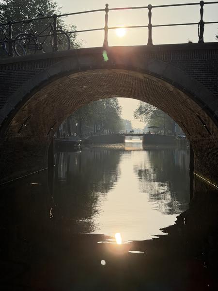 The photo captures a serene sunrise over the Herengracht canal in Centrum, Amsterdam, with the sun's golden light reflecting beautifully on the calm water
